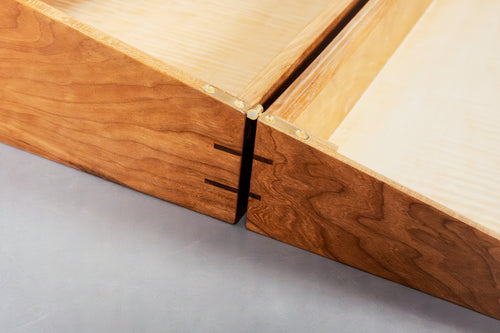 Close-up of a wooden drawer or cabinet corner showing two wood-toned sides with a black splines and brass hardware, photographed on a gray surface.