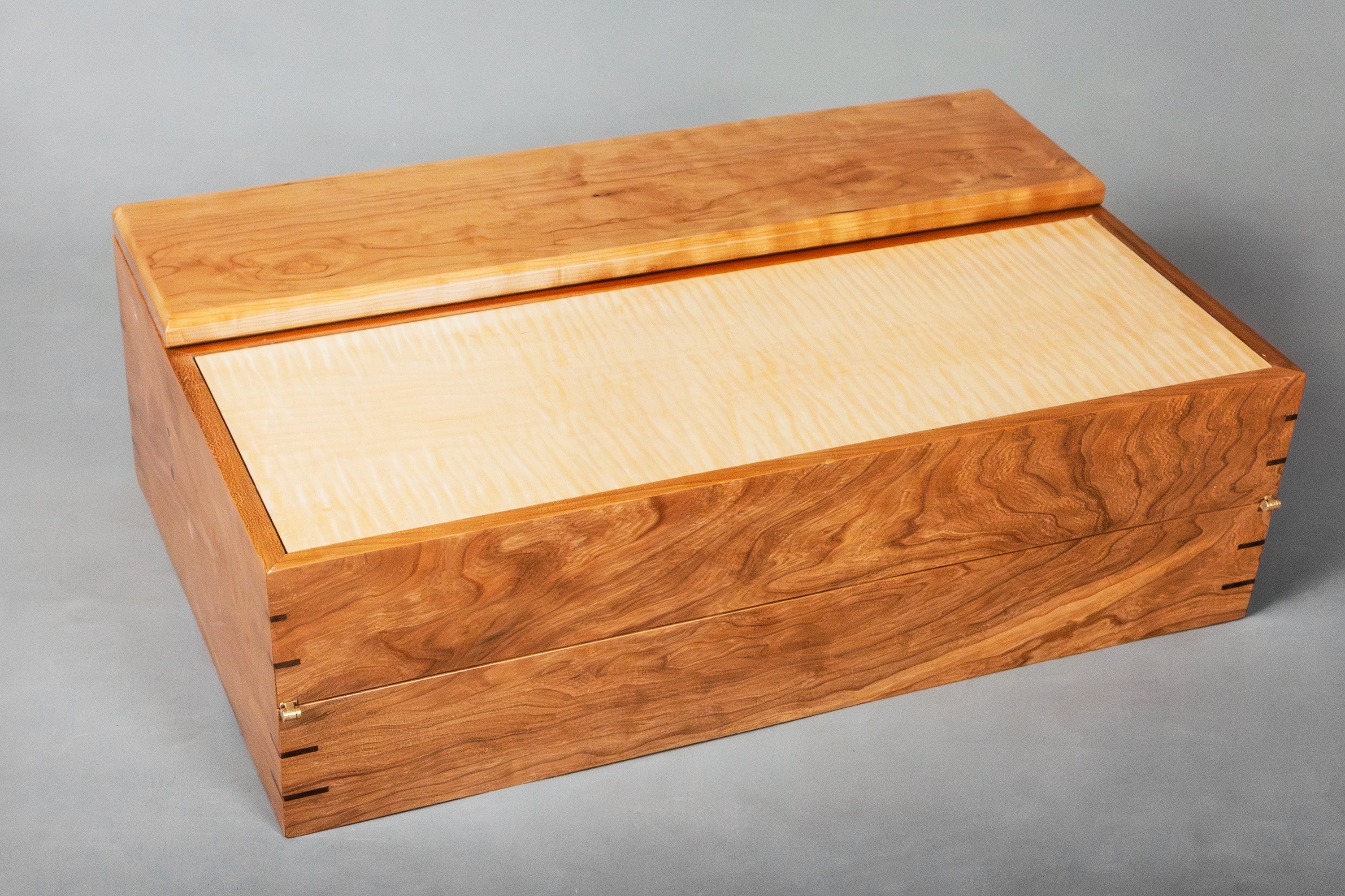A rectangular wooden writing storage box with a hinged lid, featuring a warm cherry wood grain exterior and a curly maple natural wood inner panel, photographed on a neutral gray background.